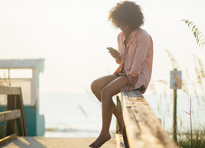 Person checking phone sitting on wooden fence at the beach.